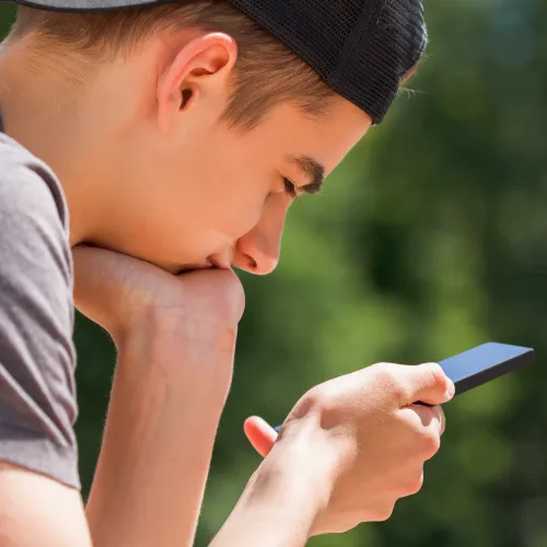 Teenager with cap on sat with head resting on left hand. Looking at black phone in right hand