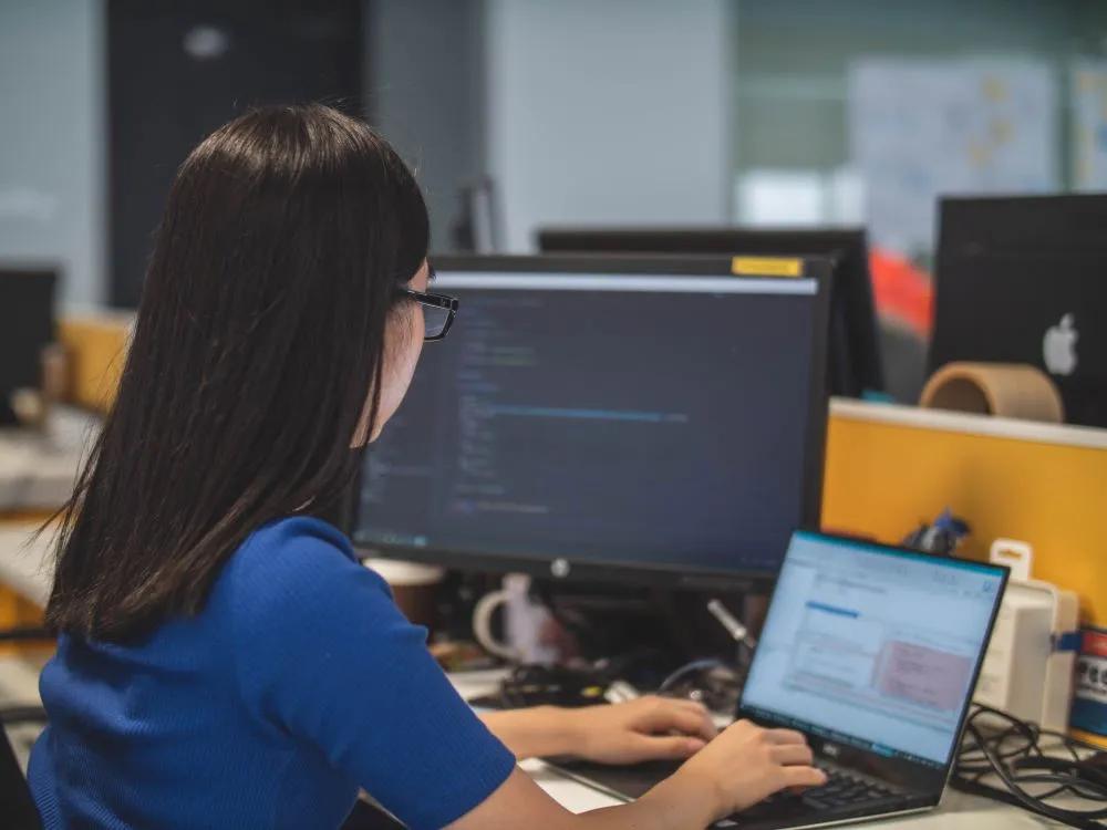 Woman coding on laptop with monitor to left
